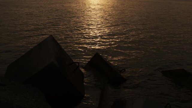 Silhouette of large concrete breakwater blocks protecting a harbor structure with golden sun reflections on the sea water surface during sunset