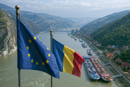 Union flag and Romanian flag flying over the Danube River in Romania's countryside with ships docked along the riverbank