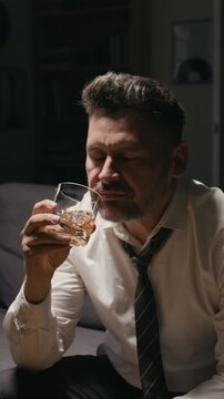 Vertical video of middle aged Caucasian businessman drinking whiskey from glass while suffering from burnout. Ideal for mental health awareness and substance abuse rehabilitation campaigns