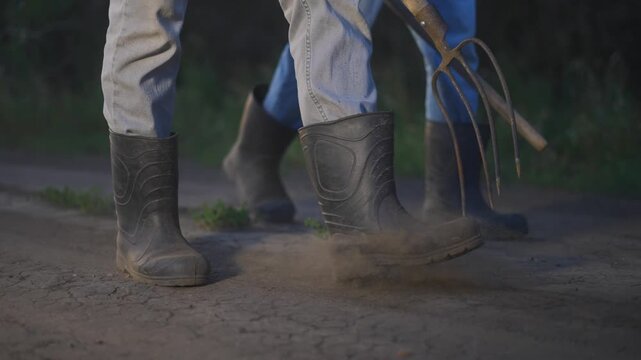 Farmer boots pitchforks road. The man and woman walk on the dirt path, using a farming fork as they prepare the soil. Their sturdy boots kick up dust on the dirt path as they work together.