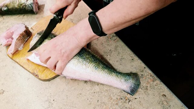 Hands of woman cutting raw fish with knife on wooden cutting board. Fresh seafood preparation at kitchen. Cooking process and fish cleaning concept.