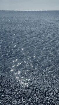 Clean transparent water on shallow sandy seashore with shells with shells on sunny summer day. Sun glare on surface of water. Small waves on sea coast. Nature backdrop. Natural background. Vertical