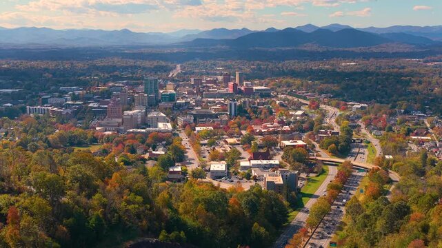 Aerial view of Asheville, North Carolina surrounded by colorful autumn forests. Downtown skyline rises against scenic Appalachian Mountains in peak fall season.
