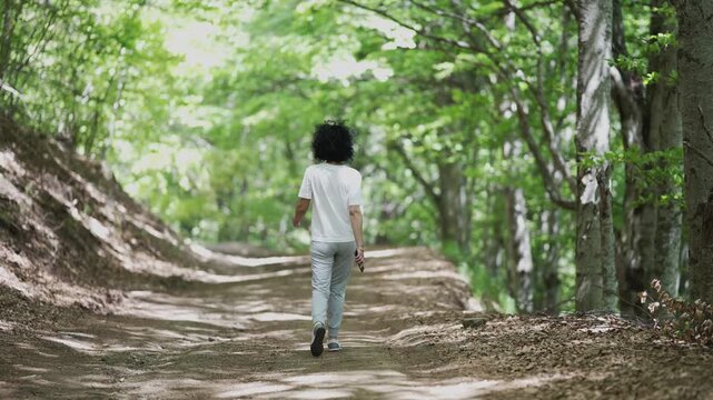 Hike woman is walking down a dirt path trail in a forest. The path is lined with trees and the woman is wearing a white shirt, slow motion