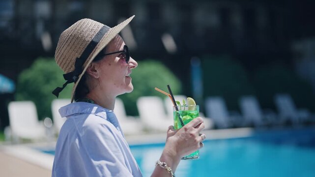 Happy smiling woman wearing a straw hat is holding a cocktail drink in a glass. The drink is green and has a straw in it. The woman is standing by a pool