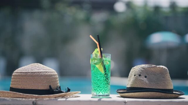A glass of green summer cocktail liquid with a straw in it sits on a swimming pool edge next to a straw hat
