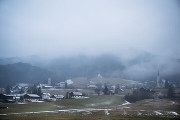 Obraz premium Wide shot of foggy alpine village with tall church spire surrounded by mist covered hills and light snow in winter. Atmospheric winter landscape in Gosau, Austria.