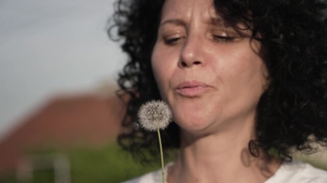A woman with curly hair is blowing on a dandelion in slow motion. The scene is peaceful and calming, as the woman takes a moment to appreciate the beauty of the flower
