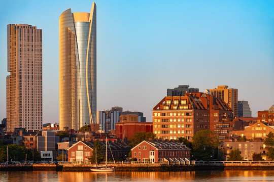 Tall skyscrapers in the financial district of Boston stand high behind the historic North End neighborhood, seen across the bay with beautiful golden sunlight reflecting on the water.
