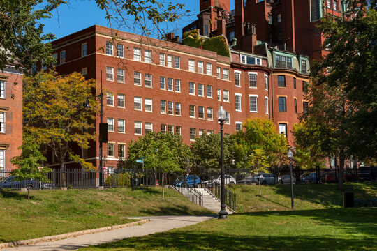Traditional red brick residential buildings of the historic Beacon Hill neighborhood surround the lush green lawn and leafy trees of Boston Common park in downtown Boston.