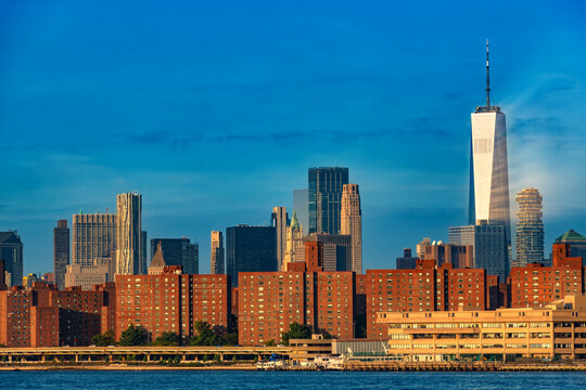 A scenic panoramic view of downtown Manhattan skyscrapers along the waterfront during early morning sunrise, with warm golden light illuminating One World Trade Center and high rise buildings.