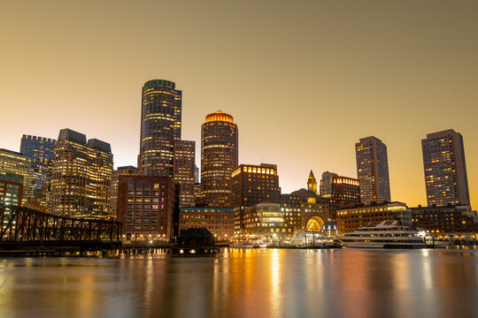 The downtown Boston skyline at sunset viewed from the vibrant Seaport District, showcasing glowing financial district buildings along the harbor waterfront.