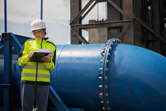 Focused professional female civil engineer holding a clipboard while inspecting large water gate infrastructure for flood control and environmental safety during an active outdoor operation.