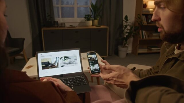 Young Caucasian couple viewed from behind browsing furniture catalog on laptop and smartphone, choosing home decor. Footage suits e-commerce advertising