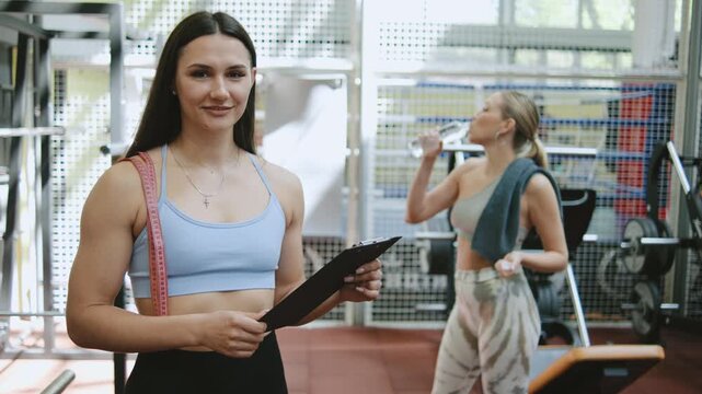 Trainer holding a clipboard stands in a gym while another person drinks water in the background