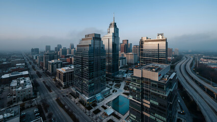 Obraz premium Aerial view of a city skyline with tall glass skyscrapers, office buildings, and a highway during a foggy morning with soft blue lighting