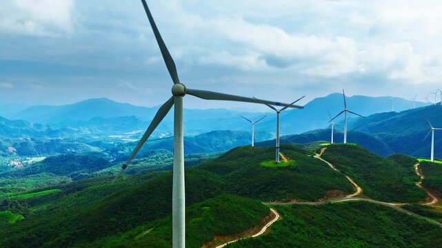 Aerial view of wind turbines on green mountain ridges, with winding dirt roads, layered peaks, and bright sunlight beneath a cloudy blue sky.