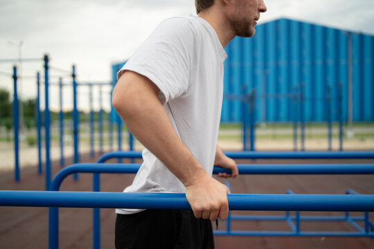 A man performs a workout on blue parallel bars outside in a city. He focuses on his routine amid the cloudy sky