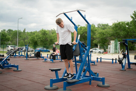 A man exercises on parallel bars in an outdoor gym while others look on. The weather is overcast and warm
