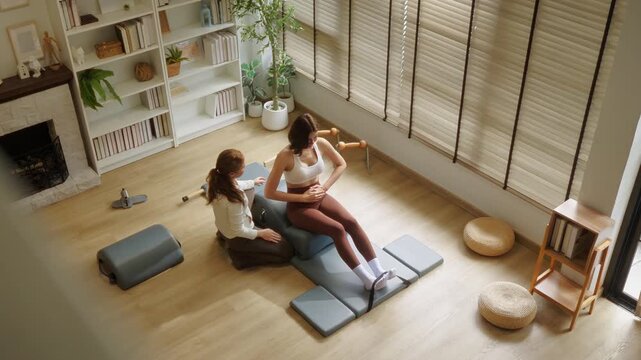 Overhead view of a personal trainer assisting a woman during a Pilates stretch on a studio mat, in a cozy sunlit room with bookshelves and green plants.	