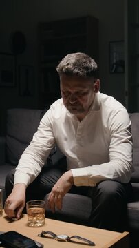 Vertical shot of depressed middle aged Caucasian man looking at wedding ring, putting it down and drinking whiskey. Footage represents divorce, depression, alcoholism and relationship crisis
