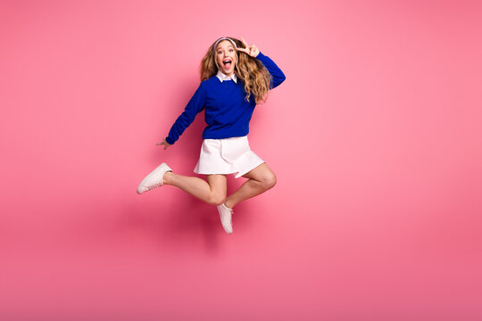 Young girl in blue sweater and white skirt jumps happily against pink background in a playful fashion scene