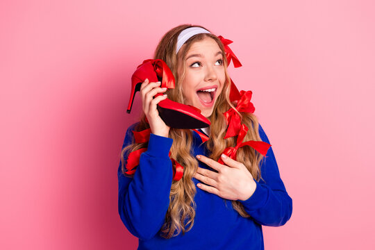 Young woman with blonde hair wears blue sweater and pink background while playfully using red high heel as phone and surrounded by ribbons