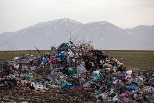 Large pile of plastic and household waste dumped in a rural agricultural area, with snow-covered mountains in the background, highlighting environmental pollution and improper waste disposal.
