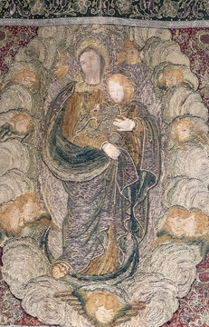 An antique embroidered textile artwork depicts the Virgin Mary holding infant Jesus among flying cherubs.