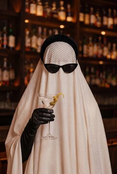 Stylish Halloween ghost holding martini cocktail with sunglasses veil and black gloves in elegant bar