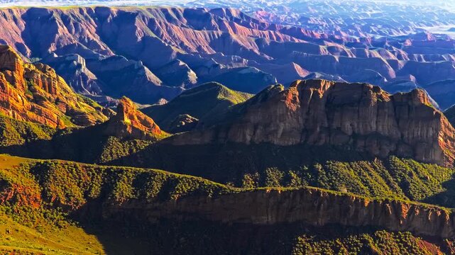 Aerial view of layered red cliffs and rugged mountain ridges with green vegetation, deep valleys, and warm sunlight across an expansive landscape.
