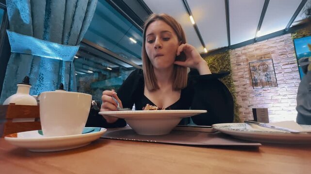Business woman is eating an organic salad meal at a restaurant. She is dressed in black has a spoon in her hand. The table is set with a plate of food and a cup. The atmosphere is casual and relaxed