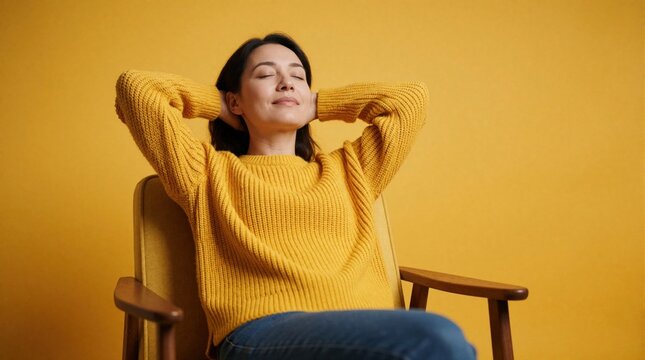 Young woman relaxes comfortably in chair with hands behind head. Smiles softly with eyes closed, enjoying moment of pure peace, quiet. Bright yellow sweater adds cheerful pop of color to simple