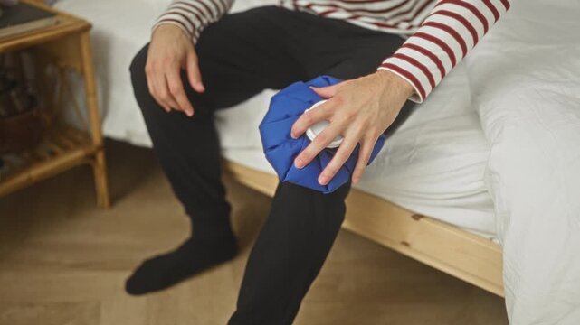 Young man holding blue ice pack to right knee while sitting on bed in building wearing striped pajamas and black socks, indicating knee pain; relief recovery.