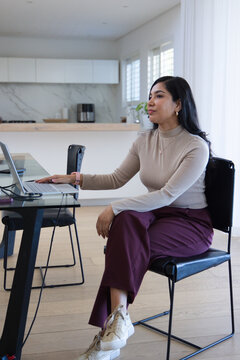 Adult woman sitting at glass dining table in kitchen in beige top touching laptop charging cable