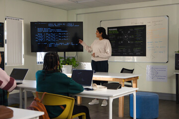 Diverse instructor teaching teenage students at room monitor showing code with laptops, copy space © WavebreakMediaMicro