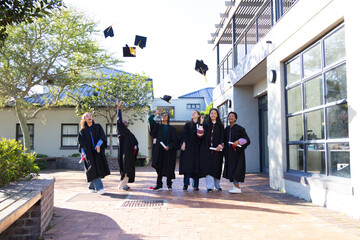 Group of six grads wearing black gowns tossing caps and holding diplomas in sunlit campus courtyard © WavebreakMediaMicro
