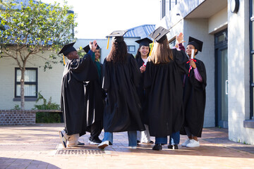 Diverse students wearing black gowns and caps, celebrating in courtyard holding red diploma tubes © WavebreakMediaMicro