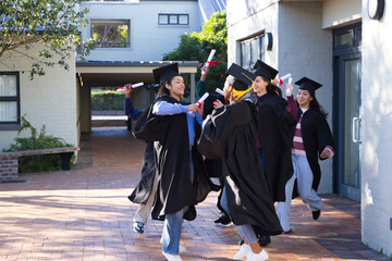 Diverse classmates celebrating on campus quad in black caps and gowns holding red-ribbon diplomas © WavebreakMediaMicro