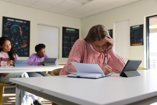 Youth female student in pink sweater writing in notebook with pen in classroom, tablet in folio