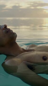 Vertical video: Floating shirtless man responding to sunset, breathing on calm sea