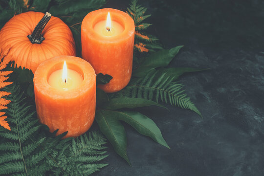 Two lit orange pillar candles with green leaves and pumpkin on a dark background.