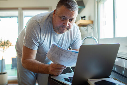 Middle-aged man reviewing bills on laptop at home kitchen counter