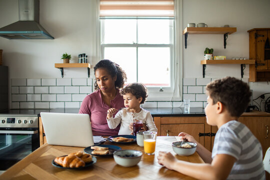 Mother working on laptop with kids at breakfast table in home kitchen