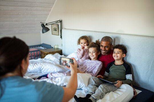 Mother photographing smiling father and kids in home bedroom