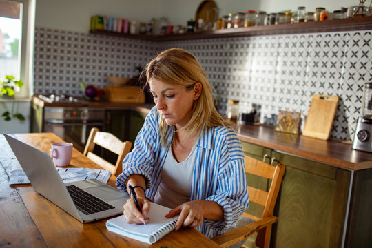 Woman taking notes while working on laptop in home kitchen