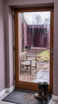 Pair of muddy rubber wellington boots standing on entryway mat beside glass door covered in raindrops. Wooden bench in wet courtyard seen through rain streaked window pane on gloomy autumn day
