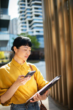 Businesswoman using smartphone with folder on modern city street
