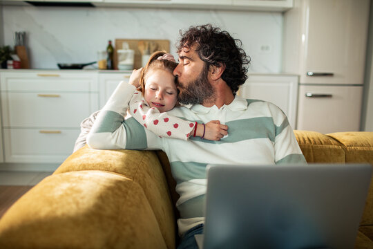 Father kissing daughter on sofa at home with laptop