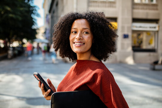 Smiling young woman with smartphone and laptop on city street